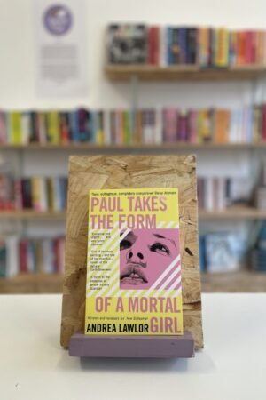 Cymraeg: Copi o 'Paul Takes the Form of a Mortal Girl' yn sefyll ar stondin llyfrau, tu blaen silffoedd o lyfrau yn y cefndir. | English: A copy of 'Paul Takes the Form of a Mortal Girl' sits on a stand in front of multiple shelves of other books.