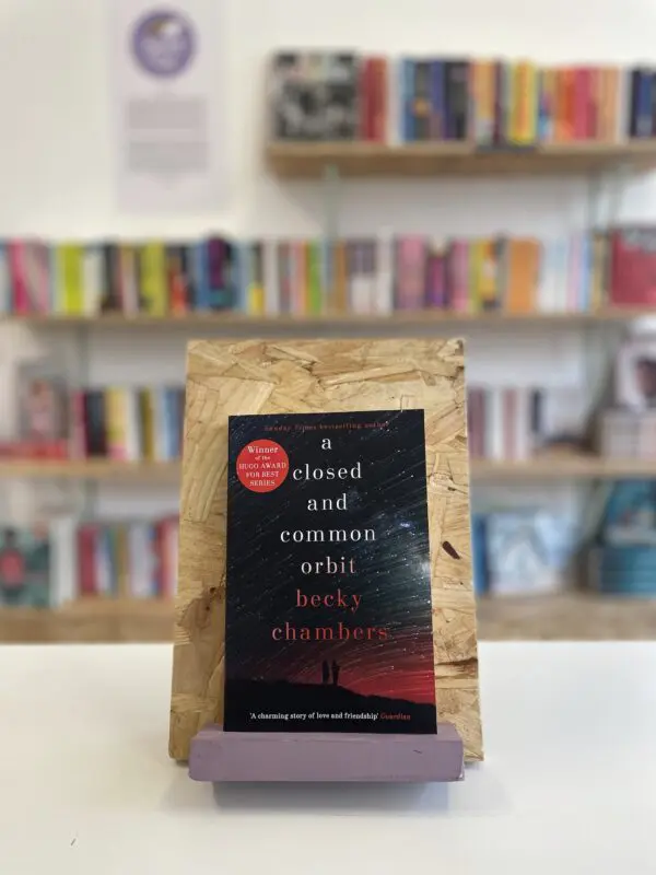 A copy of 'A Closed and Common Orbit' sits on a stand in front of multiple shelves of other books.