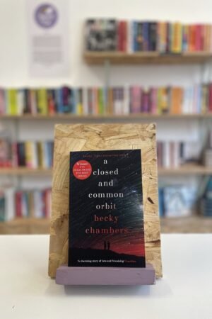 A copy of 'A Closed and Common Orbit' sits on a stand in front of multiple shelves of other books.