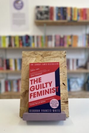 Cymraeg: Copi o 'The Guilty Feminist' yn sefyll ar stondin llyfrau, tu blaen silffoedd o lyfrau yn y cefndir. | English: A copy of 'The Guilty Feminist' sits on a stand in front of multiple shelves of other books.