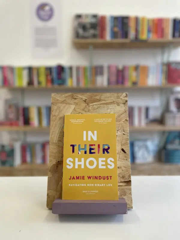 Cymraeg: Copi o 'In Their Shoes' yn sefyll ar stondin llyfrau, tu blaen silffoedd o lyfrau yn y cefndir. | English: A copy of 'In Their Shoes' sits on a stand in front of multiple shelves of other books.