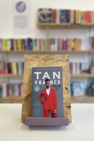 Cymraeg: Copi o 'Naturally Tan' yn sefyll ar stondin llyfrau, tu blaen silffoedd o lyfrau yn y cefndir. | English: A copy of 'Naturally Tan' sits on a stand in front of multiple shelves of other books.