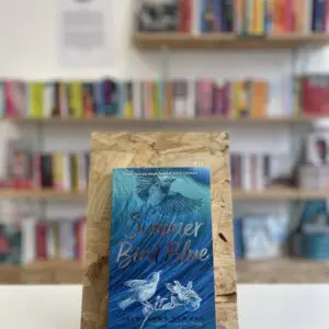 A copy of 'Summer Bird Blue' sits on a stand in front of multiple shelves of other books.