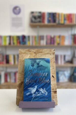 A copy of 'Summer Bird Blue' sits on a stand in front of multiple shelves of other books.