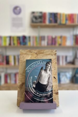 Cymraeg: Copi o 'Youngman' yn sefyll ar stondin llyfrau, tu blaen silffoedd o lyfrau yn y cefndir. | English: A copy of 'Youngman' sits on a stand in front of multiple shelves of other books.