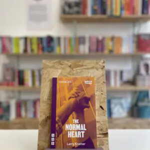 Cymraeg: Copi o 'The Normal Heart' yn sefyll ar stondin llyfrau, tu blaen silffoedd o lyfrau yn y cefndir. | English: A copy of 'The Normal Heart' sits on a stand in front of multiple shelves of other books.