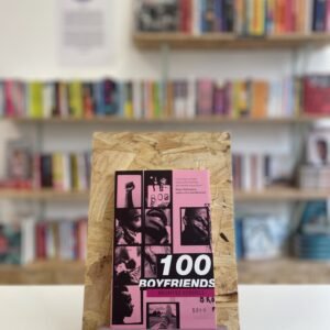 Cymraeg: Copi o '100 Boyfriends' yn sefyll ar stondin llyfrau, tu blaen silffoedd o lyfrau yn y cefndir. | English: A copy of '100 Boyfriends' sits on a stand in front of multiple shelves of other books.
