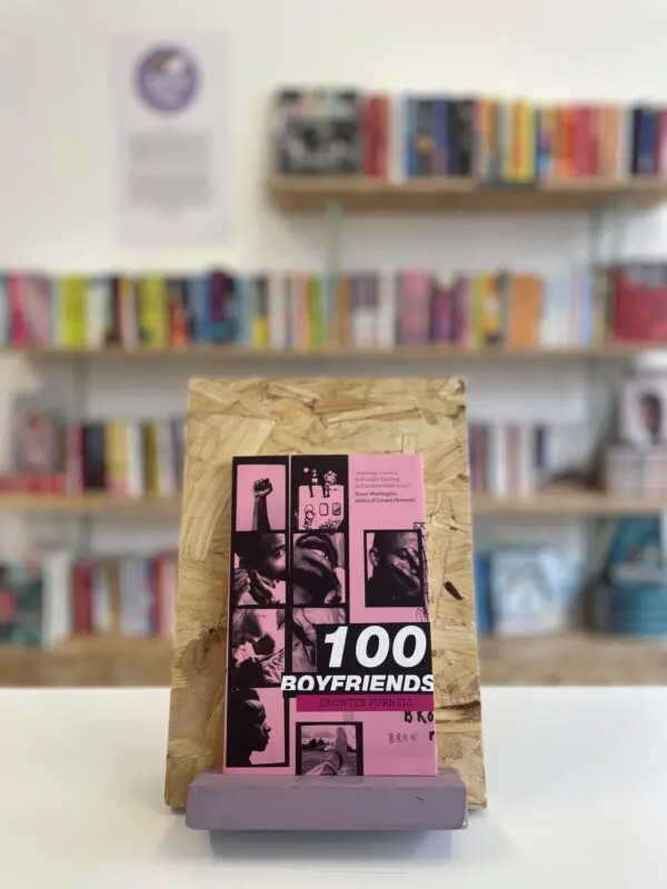 A copy of '100 Boyfriends' sits on a stand in front of multiple shelves of other books.