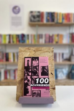 A copy of '100 Boyfriends' sits on a stand in front of multiple shelves of other books.