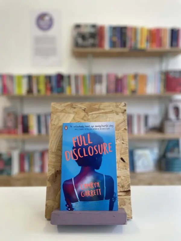 Cymraeg: Copi o 'Full Disclosure' yn sefyll ar stondin llyfrau, tu blaen silffoedd o lyfrau yn y cefndir. | English: A copy of 'Full Disclosure' sits on a stand in front of multiple shelves of other books.