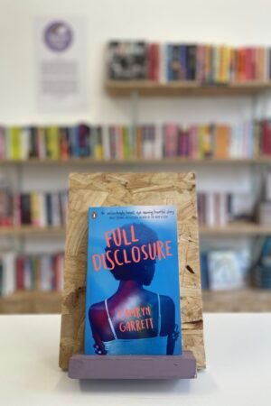 Cymraeg: Copi o 'Full Disclosure' yn sefyll ar stondin llyfrau, tu blaen silffoedd o lyfrau yn y cefndir. | English: A copy of 'Full Disclosure' sits on a stand in front of multiple shelves of other books.