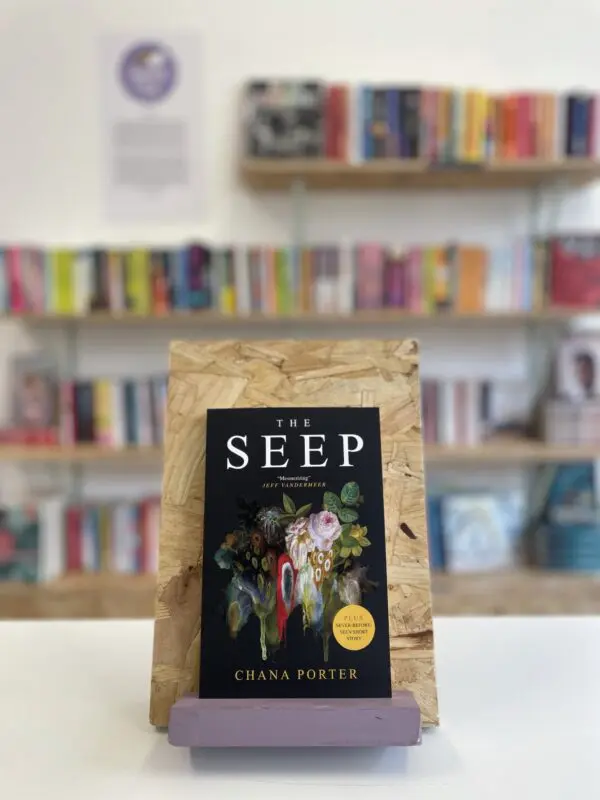 Cymraeg: Copi o 'The Seep' yn sefyll ar stondin llyfrau, tu blaen silffoedd o lyfrau yn y cefndir. | English: A copy of 'The Seep' sits on a stand in front of multiple shelves of other books.