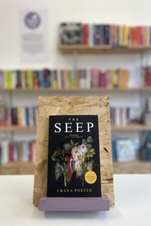 Cymraeg: Copi o 'The Seep' yn sefyll ar stondin llyfrau, tu blaen silffoedd o lyfrau yn y cefndir. | English: A copy of 'The Seep' sits on a stand in front of multiple shelves of other books.