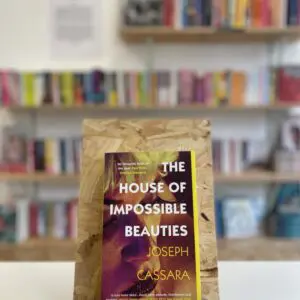 Cymraeg: Copi o 'The House of Impossible Beauties' yn sefyll ar stondin llyfrau, tu blaen silffoedd o lyfrau yn y cefndir. | English: A copy of 'The House of Impossible Beauties' sits on a stand in front of multiple shelves of other books.