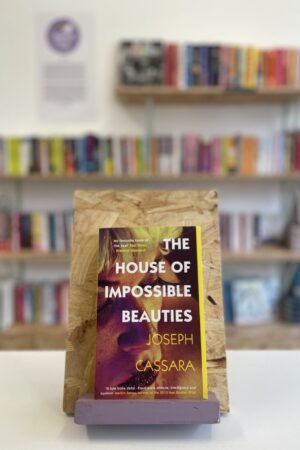 Cymraeg: Copi o 'The House of Impossible Beauties' yn sefyll ar stondin llyfrau, tu blaen silffoedd o lyfrau yn y cefndir. | English: A copy of 'The House of Impossible Beauties' sits on a stand in front of multiple shelves of other books.