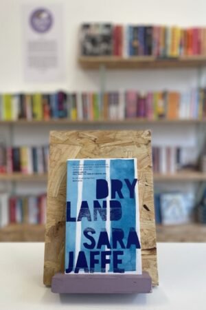 Cymraeg: Copi o 'Dryland' yn sefyll ar stondin llyfrau, tu blaen silffoedd o lyfrau yn y cefndir. | English: A copy of 'Dryland' sits on a stand in front of multiple shelves of other books.