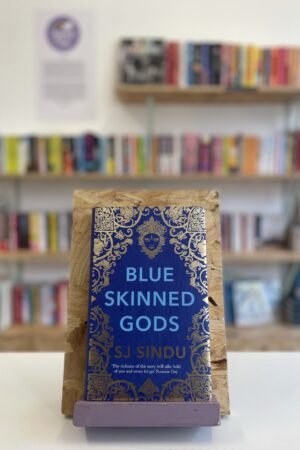 Cymraeg: Copi o 'Blue-Skinned Gods' yn sefyll ar stondin llyfrau, tu blaen silffoedd o lyfrau yn y cefndir. | English: A copy of 'Blue-Skinned Gods' sits on a stand in front of multiple shelves of other books.