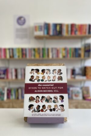 Cymraeg: Copi o 'The Essential Dykes to Watch Our For' yn sefyll ar stondin llyfrau, tu blaen silffoedd o lyfrau yn y cefndir. | English: A copy of 'The Essential Dykes to Watch Our For' sits on a stand in front of multiple shelves of other books.