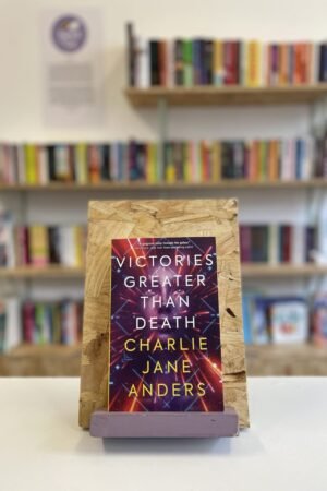 Cymraeg: Copi o 'Victories Greater Than Death' yn sefyll ar stondin llyfrau, tu blaen silffoedd o lyfrau yn y cefndir. | English: A copy of 'Victories Greater Than Death' sits on a stand in front of multiple shelves of other books.