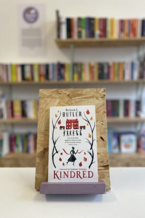 Cymraeg: Copi o 'Kindred' yn sefyll ar stondin llyfrau, tu blaen silffoedd o lyfrau yn y cefndir. | English: A copy of 'Kindred' sits on a stand in front of multiple shelves of other books.