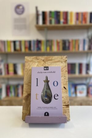 Cymraeg: Copi o 'Lote' yn sefyll ar stondin llyfrau, tu blaen silffoedd o lyfrau yn y cefndir. | English: A copy of 'Lote' sits on a stand in front of multiple shelves of other books.