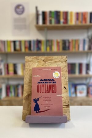 Cymraeg: Copi o 'Outlawed' yn sefyll ar stondin llyfrau, tu blaen silffoedd o lyfrau yn y cefndir. | English: A copy of 'Outlawed' sits on a stand in front of multiple shelves of other books.