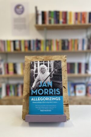 Cymraeg: Copi o 'Allegorizings' yn sefyll ar stondin llyfrau, tu blaen silffoedd o lyfrau yn y cefndir. | English: A copy of 'Allegorizings' sits on a stand in front of multiple shelves of other books.