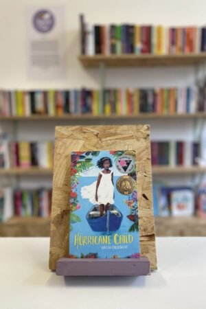 Cymraeg: Copi o 'Hurricane Child' yn sefyll ar stondin llyfrau, tu blaen silffoedd o lyfrau yn y cefndir. | English: A copy of 'Hurricane Child' sits on a stand in front of multiple shelves of other books.