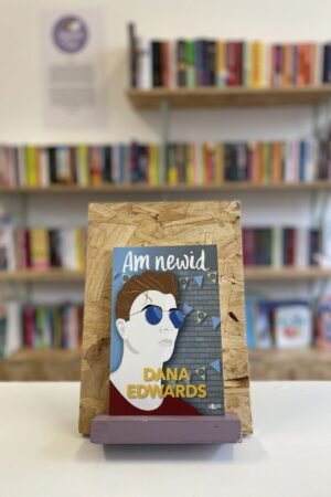 A copy of 'Am Newid' sits on a stand in front of multiple shelves of other books.