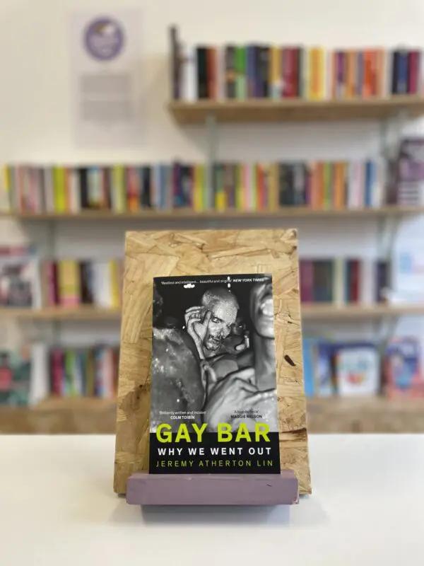 Cymraeg: Copi o 'Gay Bar' yn sefyll ar stondin llyfrau, tu blaen silffoedd o lyfrau yn y cefndir. | English: A copy of 'Gay Bar' sits on a stand in front of multiple shelves of other books.