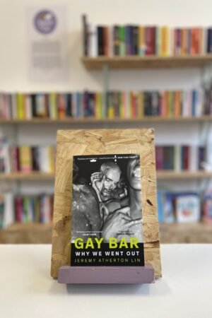 Cymraeg: Copi o 'Gay Bar' yn sefyll ar stondin llyfrau, tu blaen silffoedd o lyfrau yn y cefndir. | English: A copy of 'Gay Bar' sits on a stand in front of multiple shelves of other books.