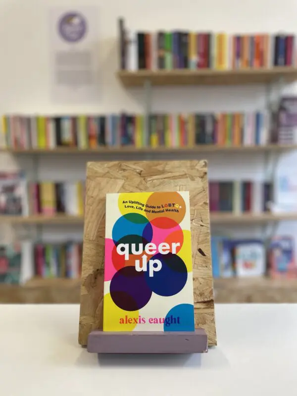 Cymraeg: Copi o 'Queer Up' yn sefyll ar stondin llyfrau, tu blaen silffoedd o lyfrau yn y cefndir. | English: A copy of 'Queer Up' sits on a stand in front of multiple shelves of other books.