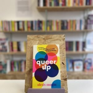 Cymraeg: Copi o 'Queer Up' yn sefyll ar stondin llyfrau, tu blaen silffoedd o lyfrau yn y cefndir. | English: A copy of 'Queer Up' sits on a stand in front of multiple shelves of other books.