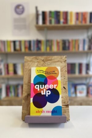 Cymraeg: Copi o 'Queer Up' yn sefyll ar stondin llyfrau, tu blaen silffoedd o lyfrau yn y cefndir. | English: A copy of 'Queer Up' sits on a stand in front of multiple shelves of other books.