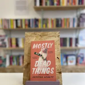 A copy of 'Mostly Dead Things' sits on a stand in front of multiple shelves of other books.