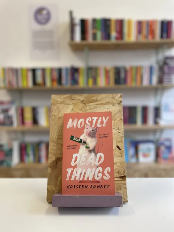 A copy of 'Mostly Dead Things' sits on a stand in front of multiple shelves of other books.