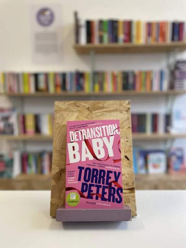 Cymraeg: Copi o 'Detransition, Baby' yn sefyll ar stondin llyfrau, tu blaen silffoedd o lyfrau yn y cefndir. | English: A copy of 'Detransition, Baby' sits on a stand in front of multiple shelves of other books.