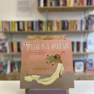 Cymraeg: Copi o 'Julian is a Mermaid' yn sefyll ar stondin llyfrau, tu blaen silffoedd o lyfrau yn y cefndir. | English: A copy of 'Julian is a Mermaid' sits on a stand in front of multiple shelves of other books.