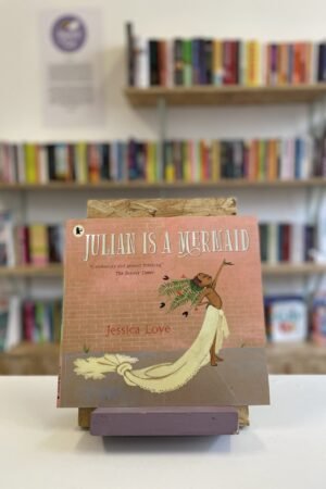 Cymraeg: Copi o 'Julian is a Mermaid' yn sefyll ar stondin llyfrau, tu blaen silffoedd o lyfrau yn y cefndir. | English: A copy of 'Julian is a Mermaid' sits on a stand in front of multiple shelves of other books.