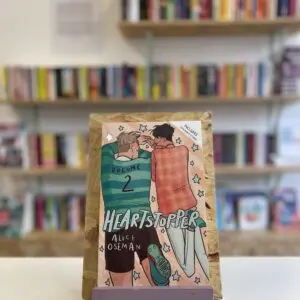 Cymraeg: Copi o 'Heartstopper vol. 2' yn sefyll ar stondin llyfrau, tu blaen silffoedd o lyfrau yn y cefndir. | English: A copy of 'Heartstopper vol. 2' sits on a stand in front of multiple shelves of other books.