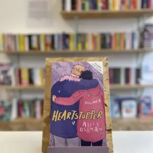 Cymraeg: Copi o 'Heartstopper vol. 4' yn sefyll ar stondin llyfrau, tu blaen silffoedd o lyfrau yn y cefndir. | English: A copy of 'Heartstopper vol. 4' sits on a stand in front of multiple shelves of other books.