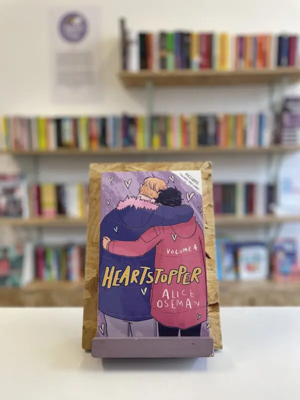 Cymraeg: Copi o 'Heartstopper vol. 4' yn sefyll ar stondin llyfrau, tu blaen silffoedd o lyfrau yn y cefndir. | English: A copy of 'Heartstopper vol. 4' sits on a stand in front of multiple shelves of other books.