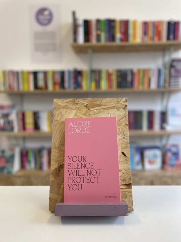 A copy of 'Your Silence Will Not Protect You' sits on a stand in front of multiple shelves of other books.