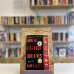 Cymraeg: Copi o 'How We Fight For Our Lives' yn sefyll ar stondin llyfrau, tu blaen silffoedd o lyfrau yn y cefndir. | English: A copy of 'How We Fight For Our Lives' sits on a stand in front of multiple shelves of other books.