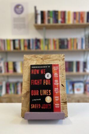 Cymraeg: Copi o 'How We Fight For Our Lives' yn sefyll ar stondin llyfrau, tu blaen silffoedd o lyfrau yn y cefndir. | English: A copy of 'How We Fight For Our Lives' sits on a stand in front of multiple shelves of other books.