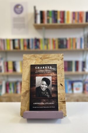 Cymraeg: Copi o 'Charred' yn sefyll ar stondin llyfrau, tu blaen silffoedd o lyfrau yn y cefndir. | English: A copy of 'Charred' sits on a stand in front of multiple shelves of other books.