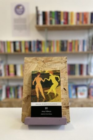 Cymraeg: Copi o 'Jill' yn sefyll ar stondin llyfrau, tu blaen silffoedd o lyfrau yn y cefndir. | English: A copy of 'Jill' sits on a stand in front of multiple shelves of other books.