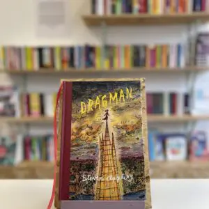 Cymraeg: Copi o 'Dragman' yn sefyll ar stondin llyfrau, tu blaen silffoedd o lyfrau yn y cefndir. | English: A copy of 'Dragman' sits on a stand in front of multiple shelves of other books.