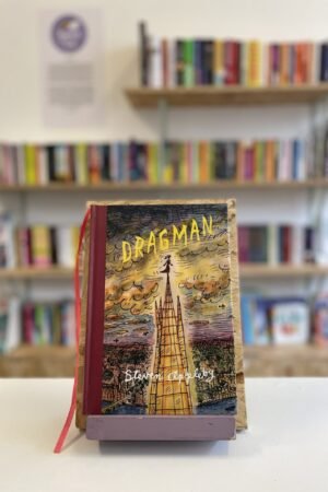 Cymraeg: Copi o 'Dragman' yn sefyll ar stondin llyfrau, tu blaen silffoedd o lyfrau yn y cefndir. | English: A copy of 'Dragman' sits on a stand in front of multiple shelves of other books.