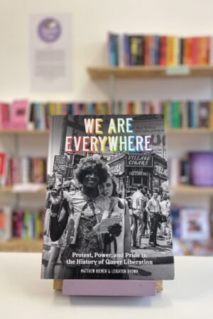 Cymraeg: Copi o 'We Are Everywhere' yn sefyll ar stondin llyfrau, tu blaen silffoedd o lyfrau yn y cefndir. | English: A copy of 'We Are Everywhere' sits on a stand in front of multiple shelves of other books.